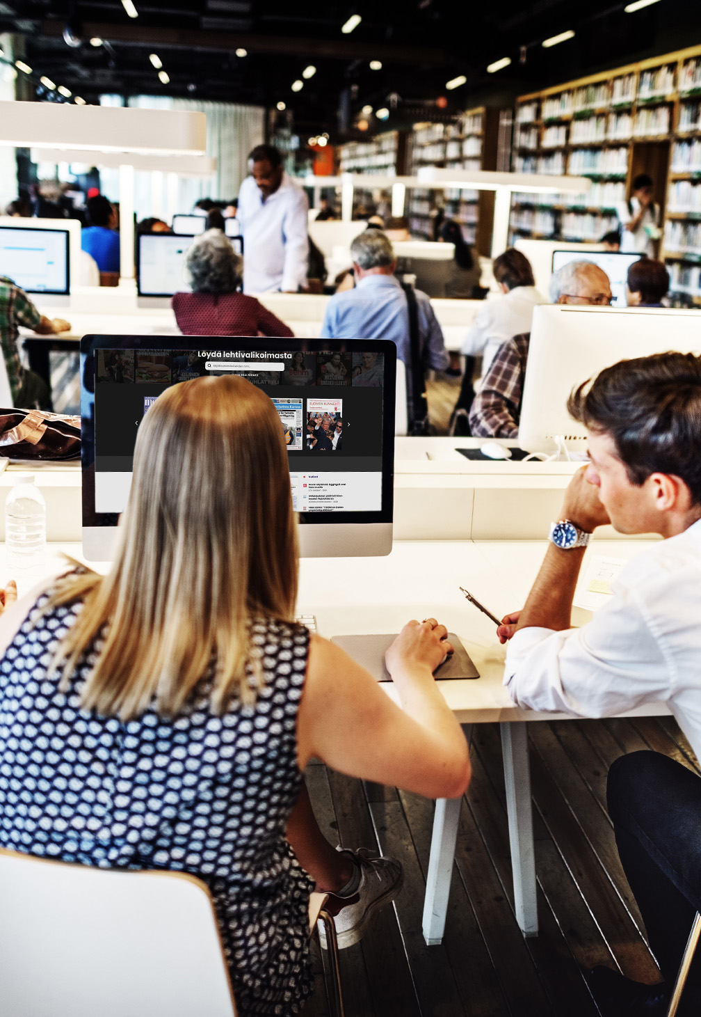 Bild som visar en biblioteksmiljö med personer som arbetar vid stationära datorer. I förgrunden ses en kvinna och en man som diskuterar innehållet på en datorskärm, vilket förstärker en samarbetsinriktad stämning.